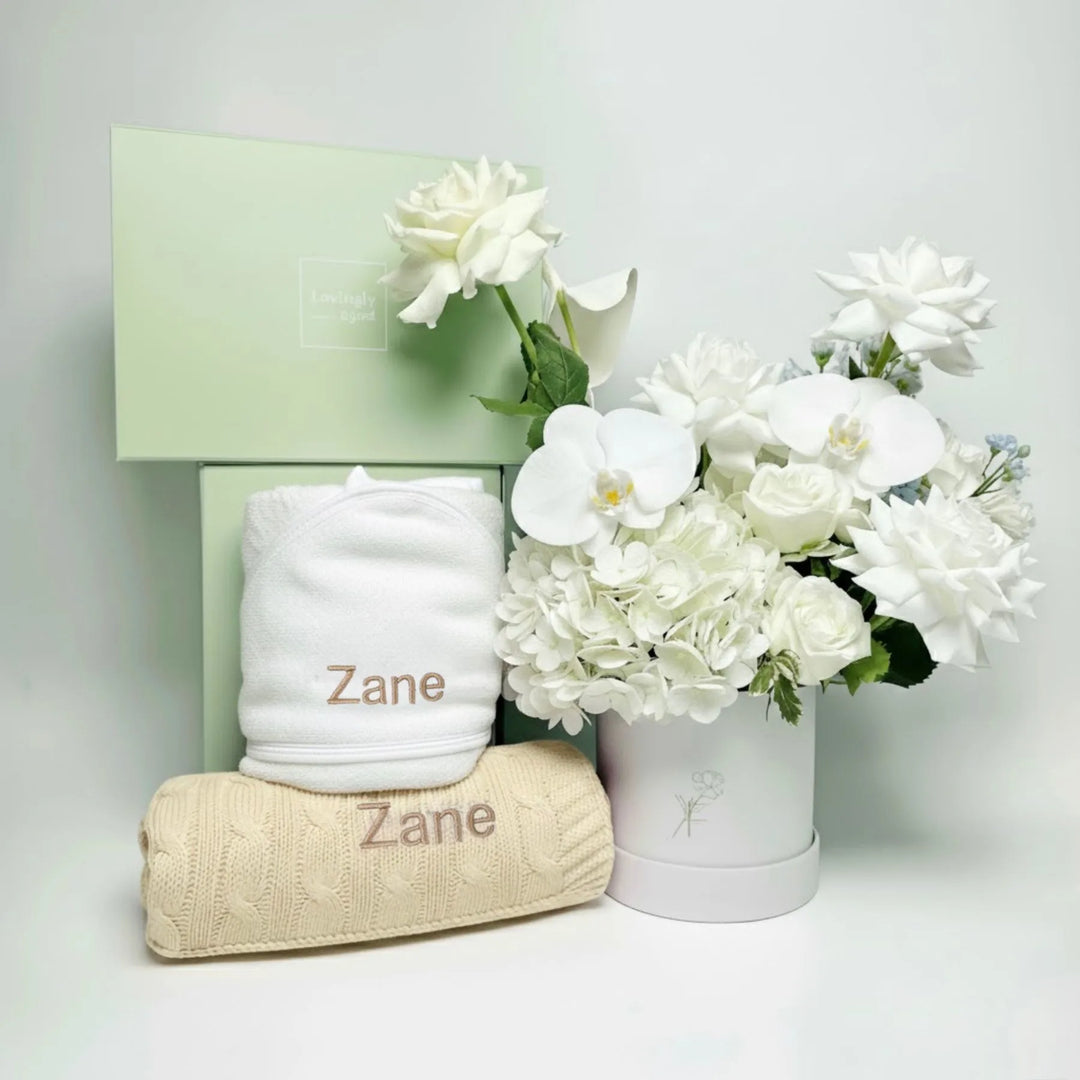 White towel and cream blanket with 'Zane' personalised, and a white flower arrangement on a light gray background.