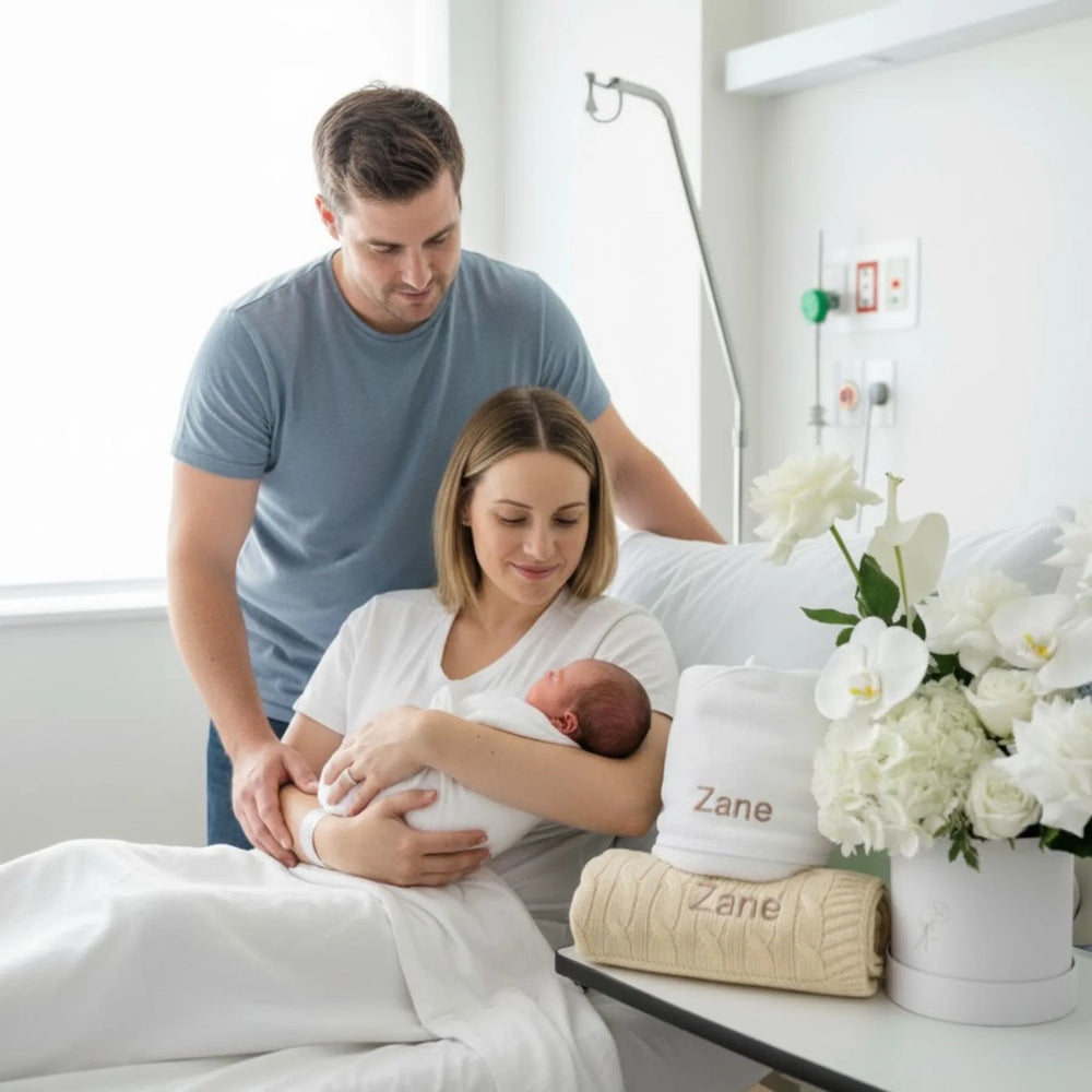 New parents with their newborn baby in a hospital room , surrounded by white flowers and personalised gifts