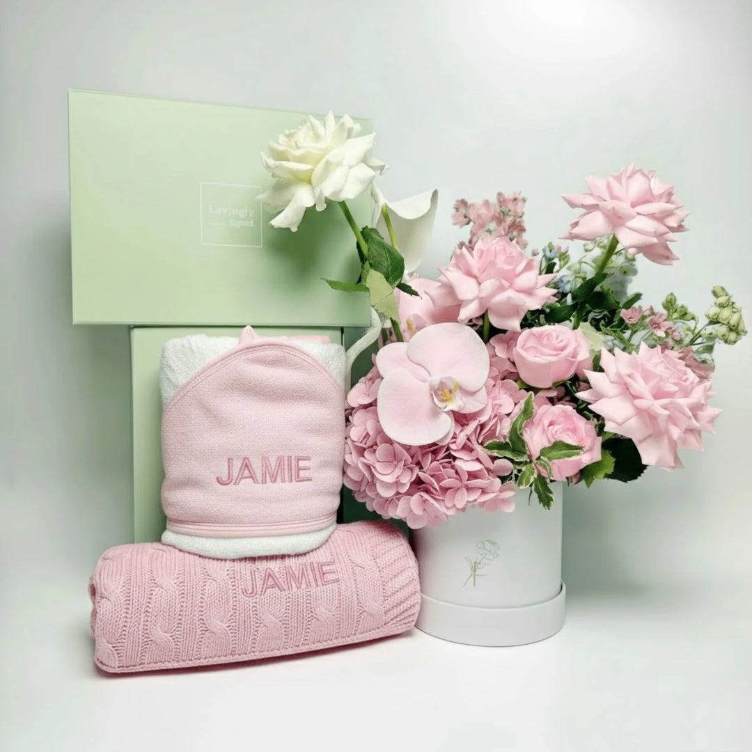 Pink baby towel and blanket with 'JAMIE' embroidery next to a bouquet of pink flowers in a white box on a light gray background.