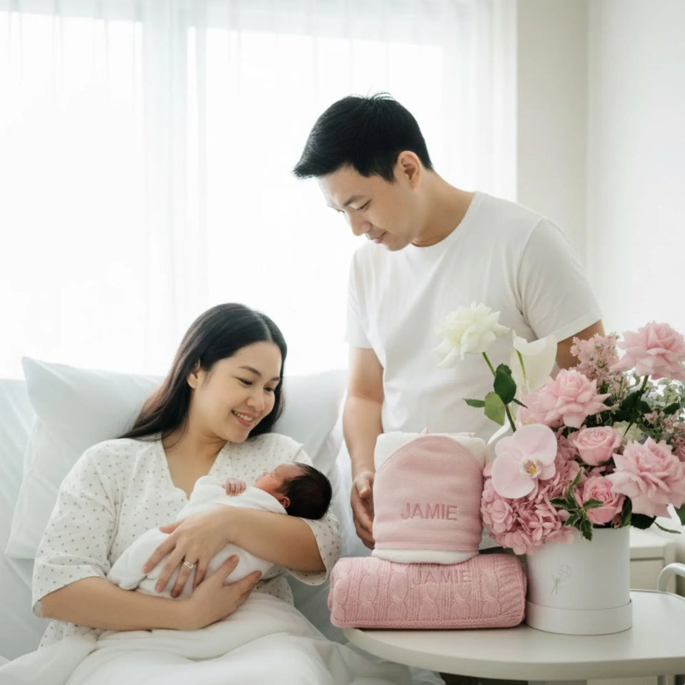 Newborn baby being held by a mum and dad standing beside them, surrounded by pink flowers, personalised blanket and towel  on a white couch.