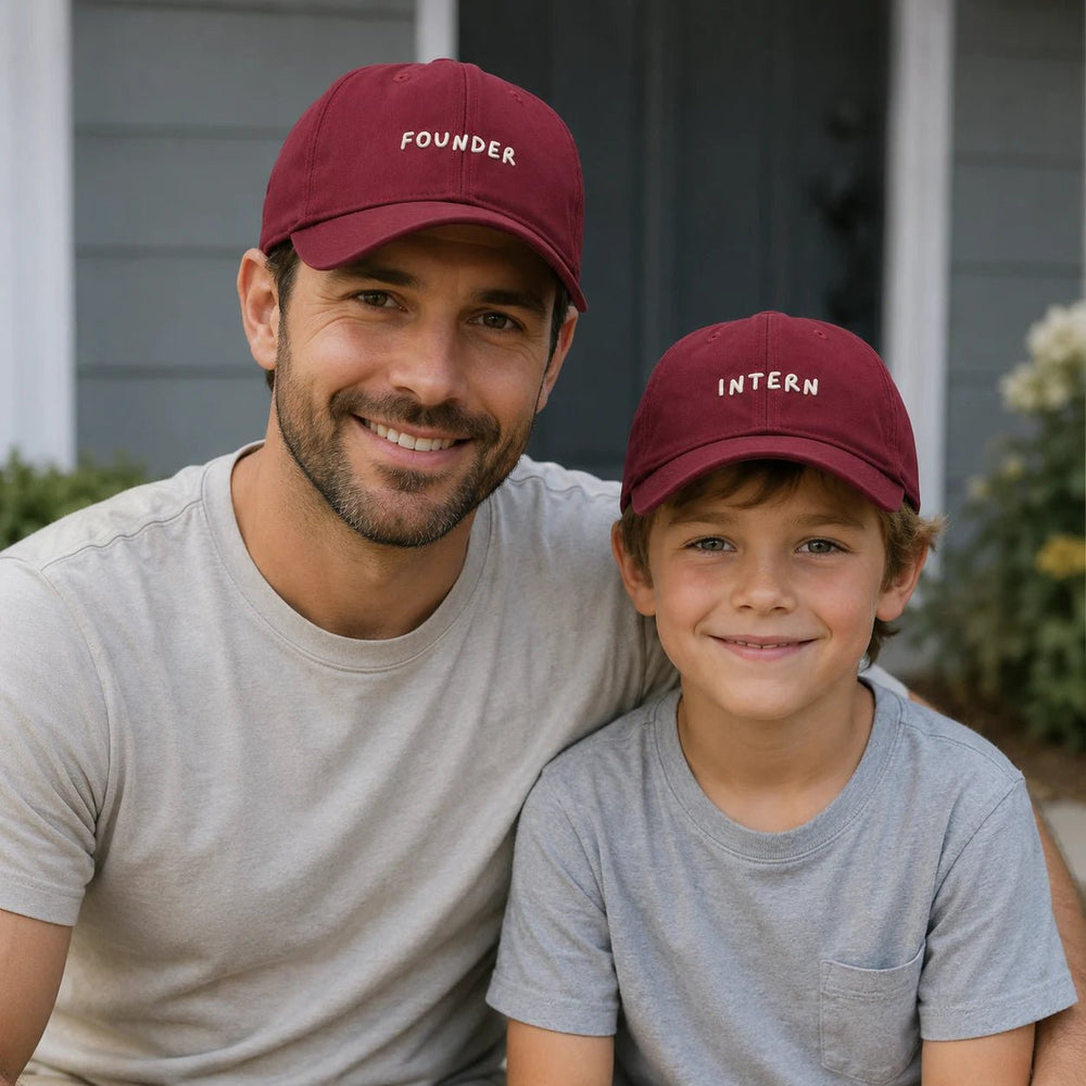 Man and child wearing maroon caps with 'Founder' and 'Intern' text, sitting outdoors.