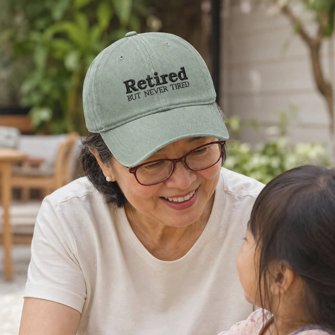 Asian grandmother wearing a “Retired But Never Tired” cap while smiling at her grandchild outdoors.