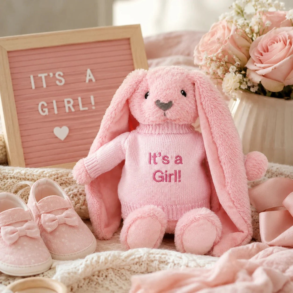 Pink bunny soft toy wearing an “It’s a Girl!” jumper, styled in a baby girl gender reveal setup with pink baby shoes, flowers, ribbon, and a letter board.