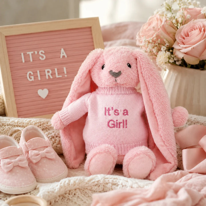 Pink bunny soft toy wearing an “It’s a Girl!” jumper, styled in a baby girl gender reveal setup with pink baby shoes, flowers, ribbon, and a letter board.