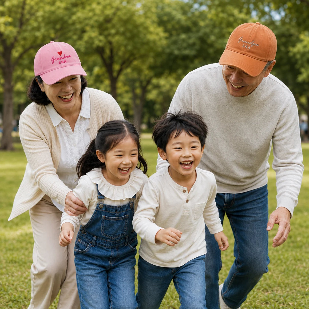 Asian grandma and grandpa wearing personalised “Grandma Era” and “Grandpa Era” caps while playing outdoors with their grandchildren in a park.
