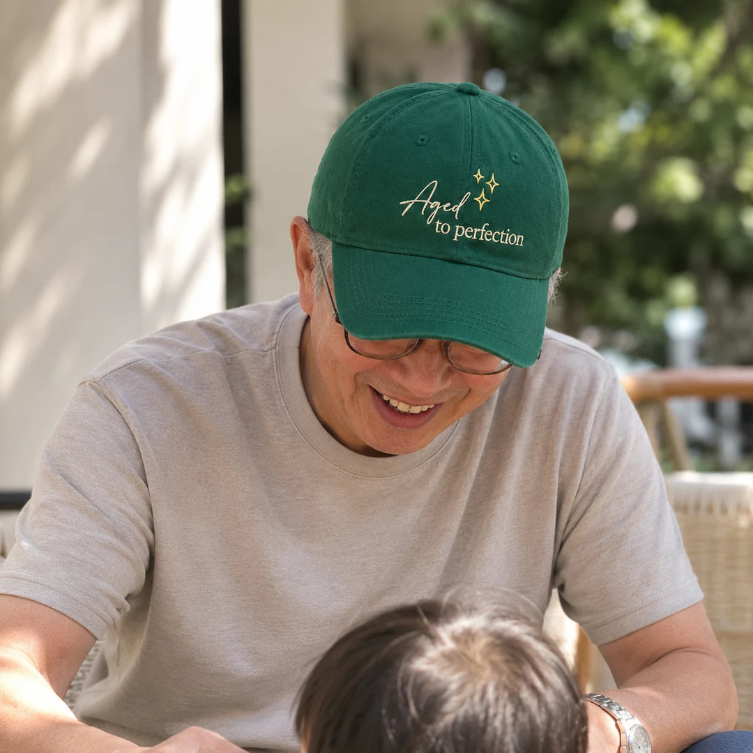 Asian grandfather wearing an “Aged to Perfection” cap while spending time with his grandchild outdoors.