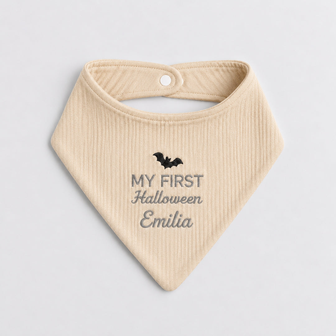 Personalised beige baby bandana bib featuring a black embroidered bat, grey “My First Halloween” lettering and the name Emilia in cursive embroidery.
