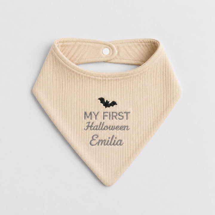 Personalised beige baby bandana bib featuring a black embroidered bat, grey “My First Halloween” lettering and the name Emilia in cursive embroidery.