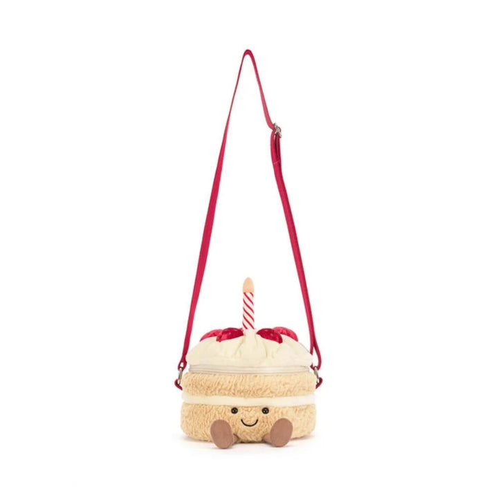 Birthday cake bag shown with the long red crossbody strap fully extended, featuring a smiling plush cake design topped with cream frosting, red berries and a striped candle.