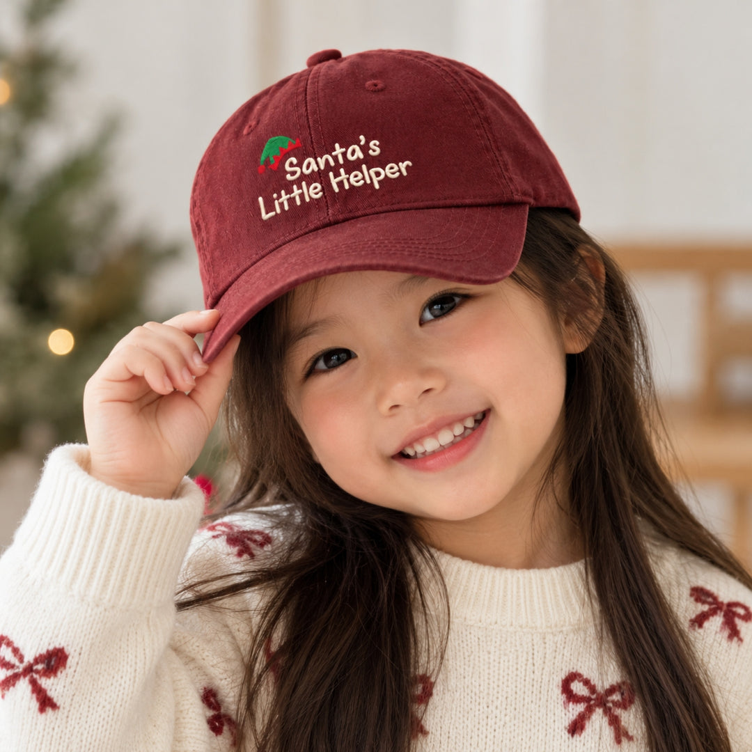 Smiling young Asian girl wearing a burgundy “Santa's Little Helper” embroidered Christmas cap and cream festive sweater, with a softly blurred Christmas tree in the background.
