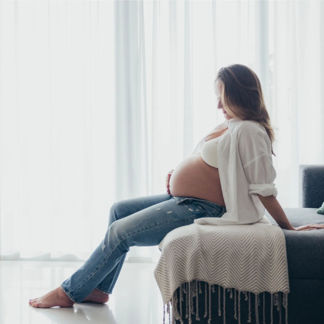 Pregnant woman sitting on a couch in a bright room with white curtains, photographed by Katie Martin-Sperry Photography