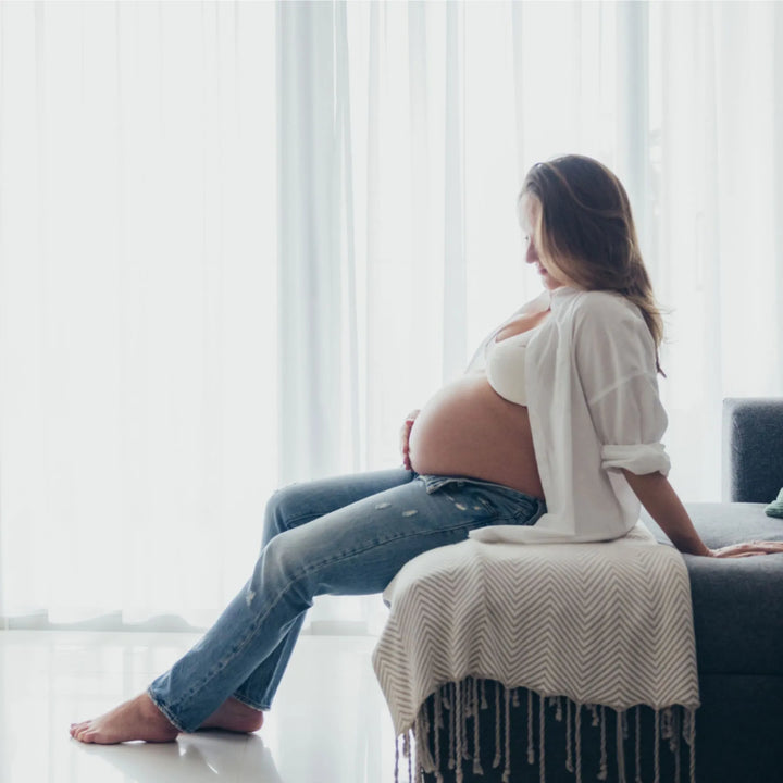 Pregnant woman sitting on a couch in a bright room with white curtains, photographed by Katie Martin-Sperry Photography