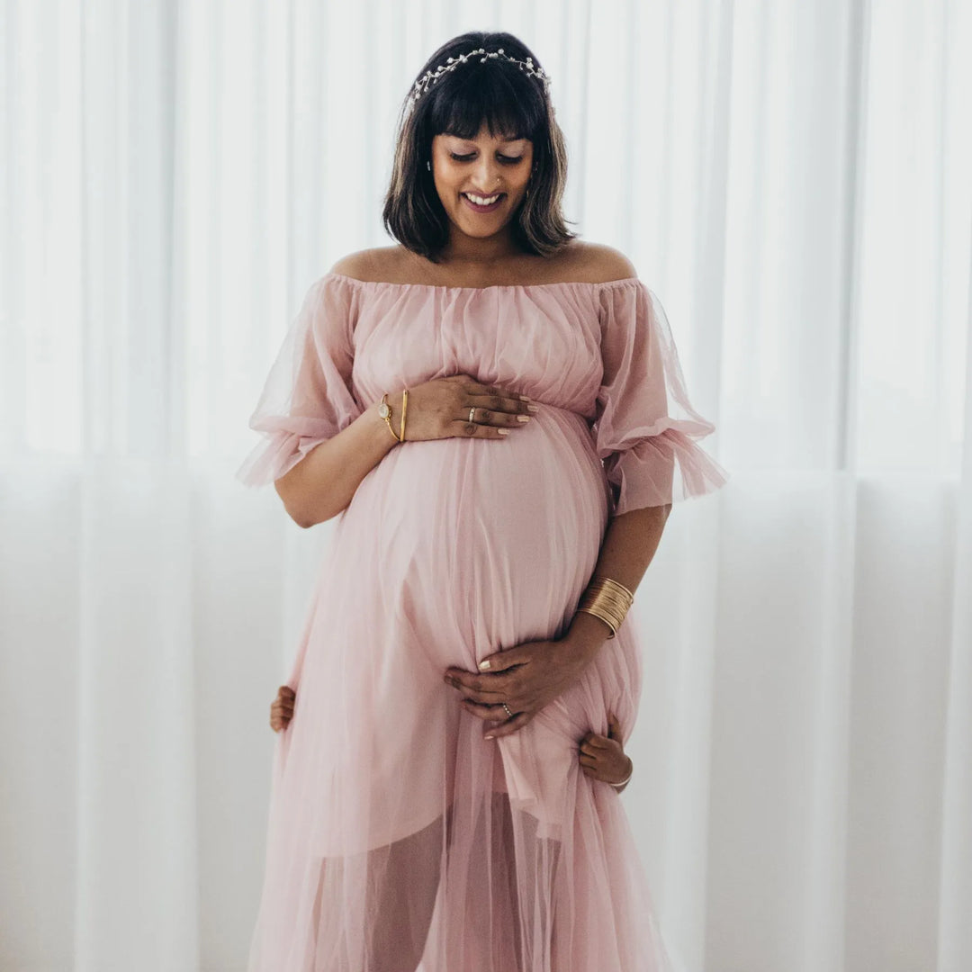 Pregnant woman in a pink dress holding her belly against a white curtain background, a beautiful maternity shoot photographed by Katie Martin-Sperry Photography