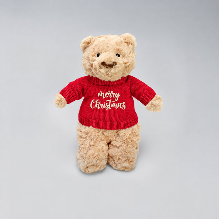 Teddy bear wearing a red knitted Christmas jumper embroidered with “Merry Christmas” in cream, photographed against a light grey studio background.