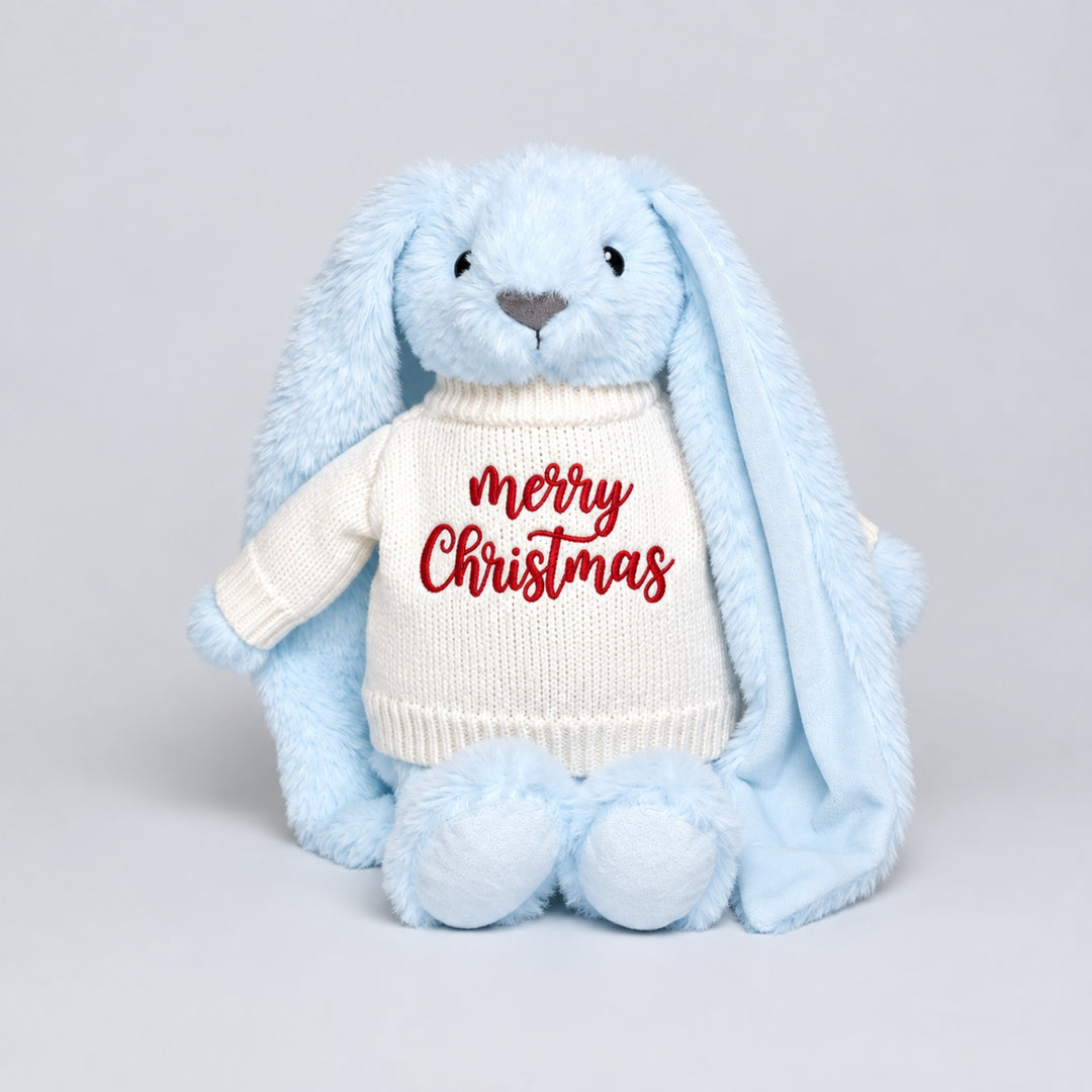 Blue plush bunny with long floppy ears wearing a cream knitted jumper embroidered with “Merry Christmas” in red, photographed against a light grey studio background.