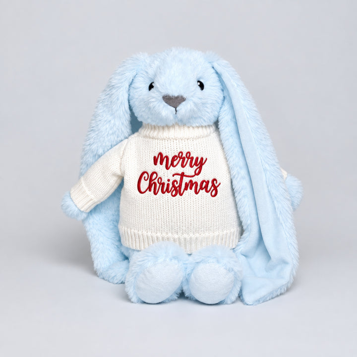 Blue plush bunny with long floppy ears wearing a cream knitted jumper embroidered with “Merry Christmas” in red, photographed against a light grey studio background.