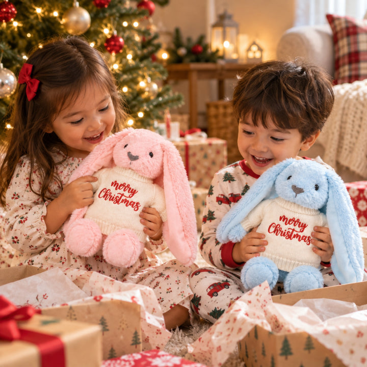 Smiling young siblings opening Christmas gifts and holding their pink and blue Merry Christmas bunnies beside a decorated Christmas tree.