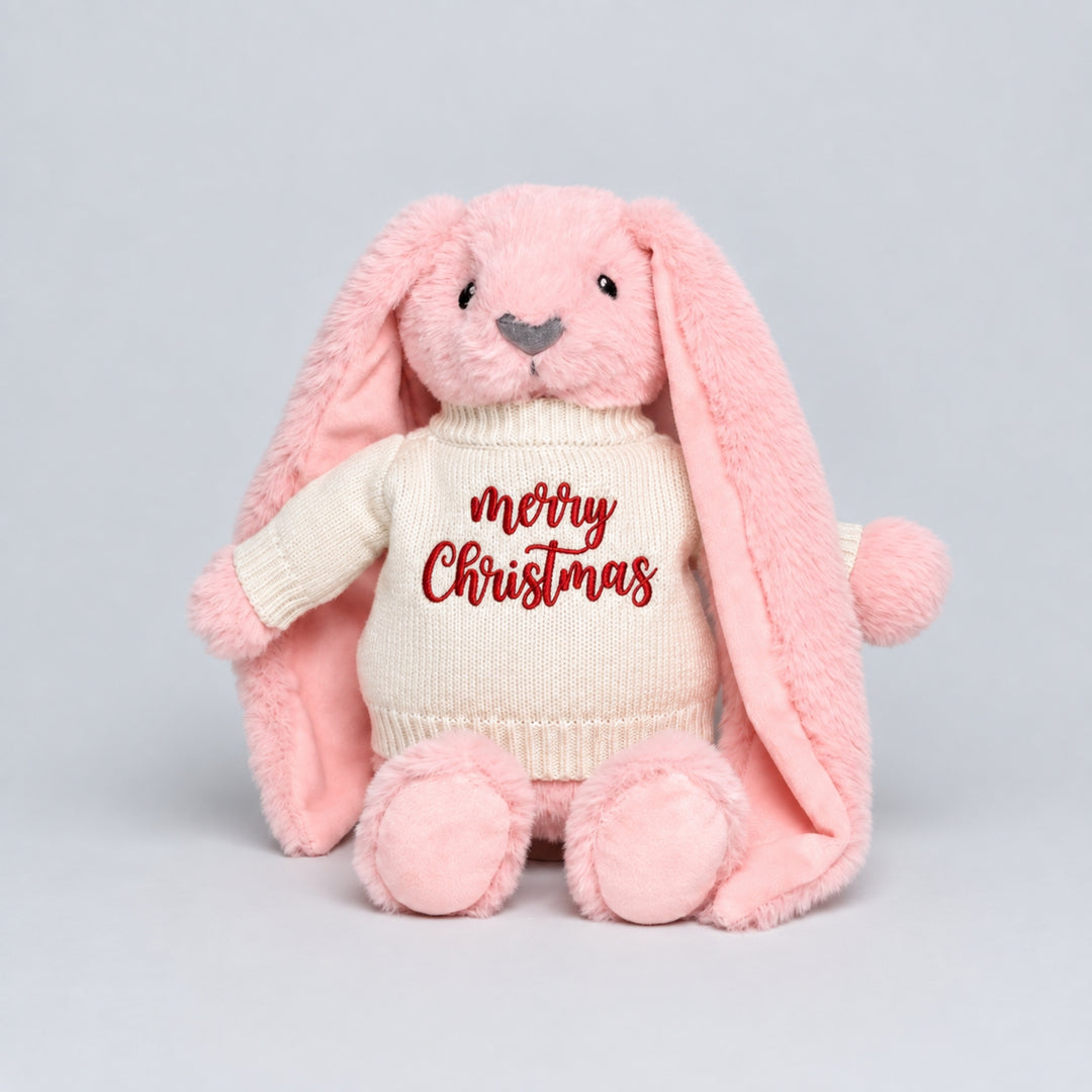 Pink plush bunny wearing a cream knitted jumper embroidered with “Merry Christmas” in red, photographed against a light grey studio background.