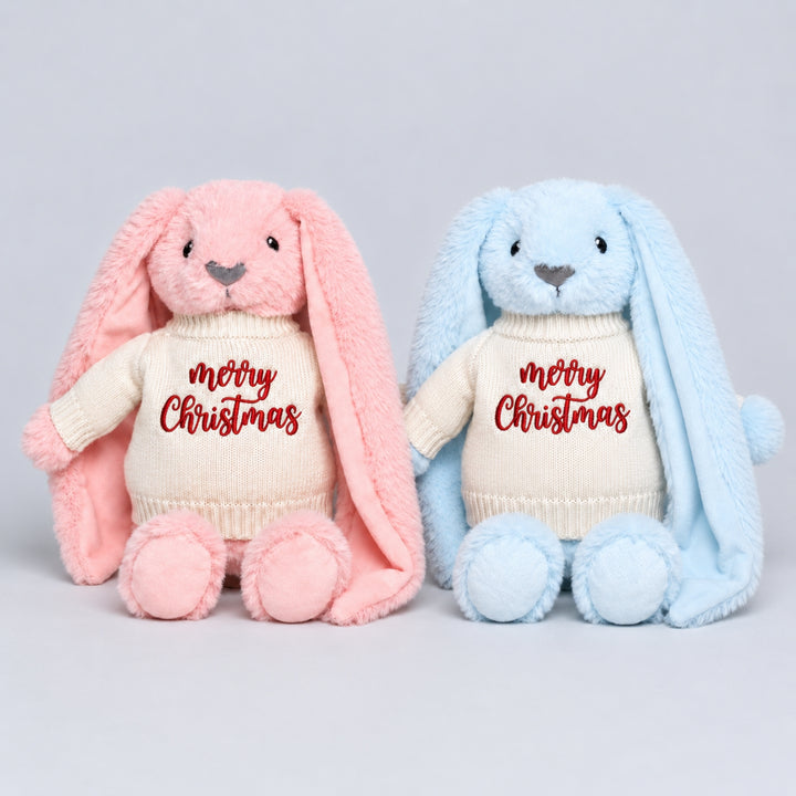 Pink and blue plush bunnies sitting side by side, each wearing a cream knitted jumper embroidered with “Merry Christmas” in red.