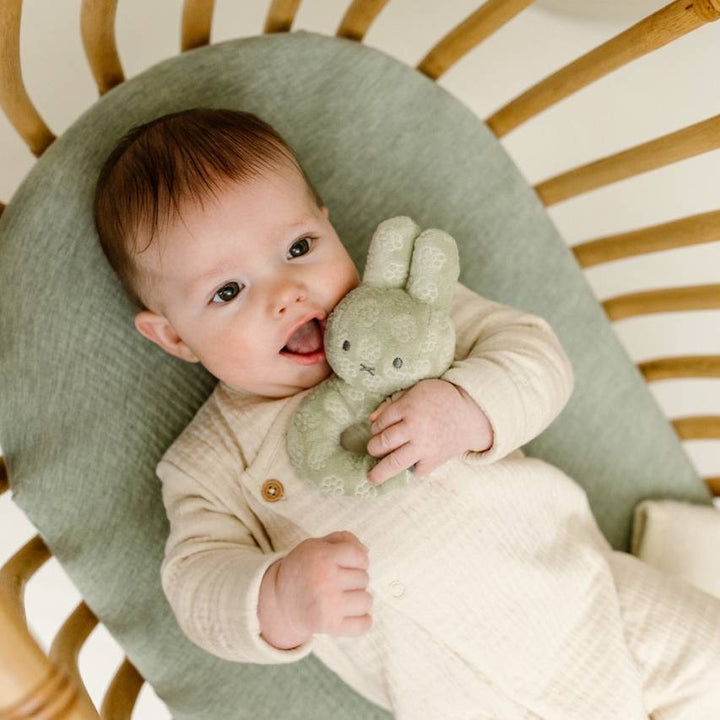 Baby holding a green miffy flower rattle in a crib with a wooden frame