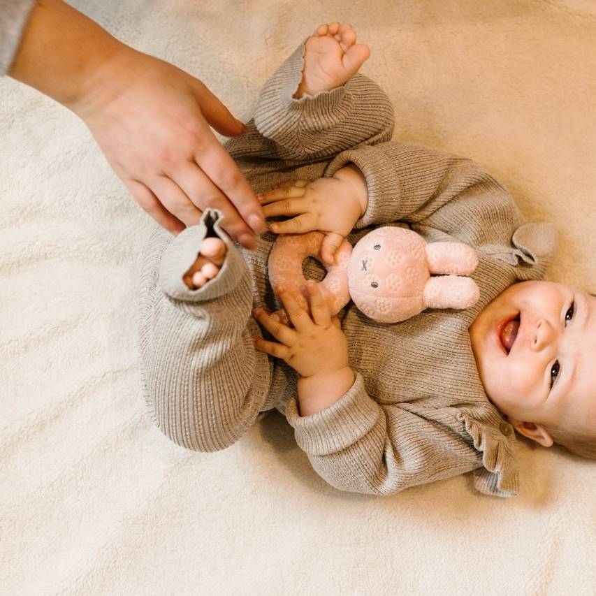 Baby lying on a soft surface with miffy pink flower rattle, wearing a cozy outfit.