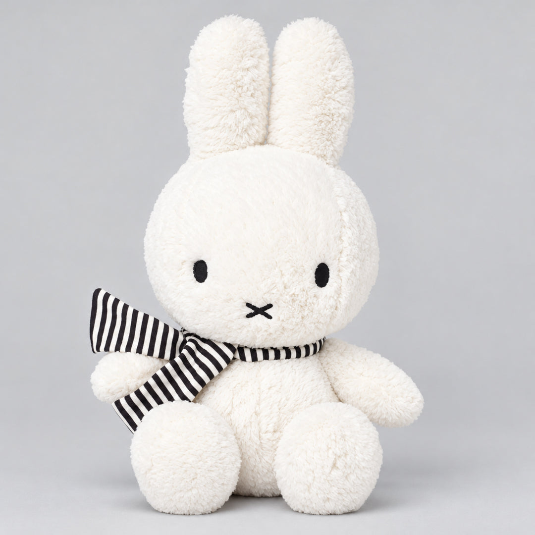 White Miffy bunny plush toy with long upright ears and a black and white striped scarf, seated against a light grey studio background.