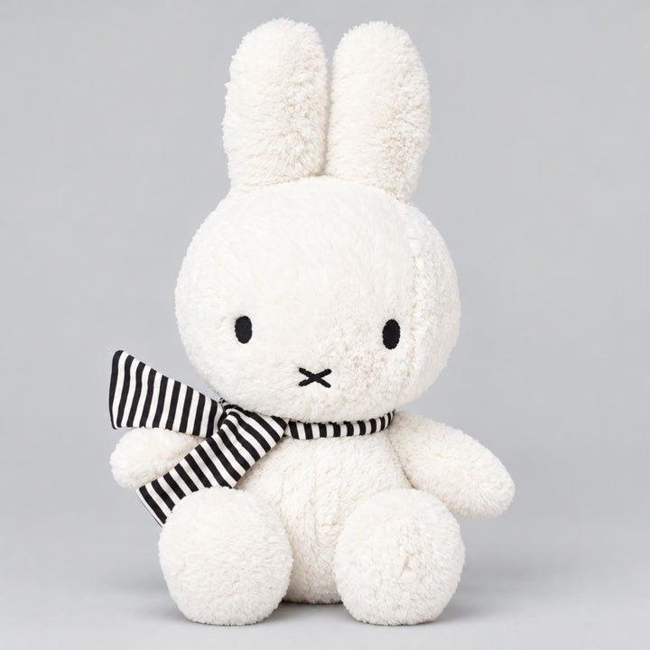 White Miffy bunny plush toy with long upright ears and a black and white striped scarf, seated against a light grey studio background.