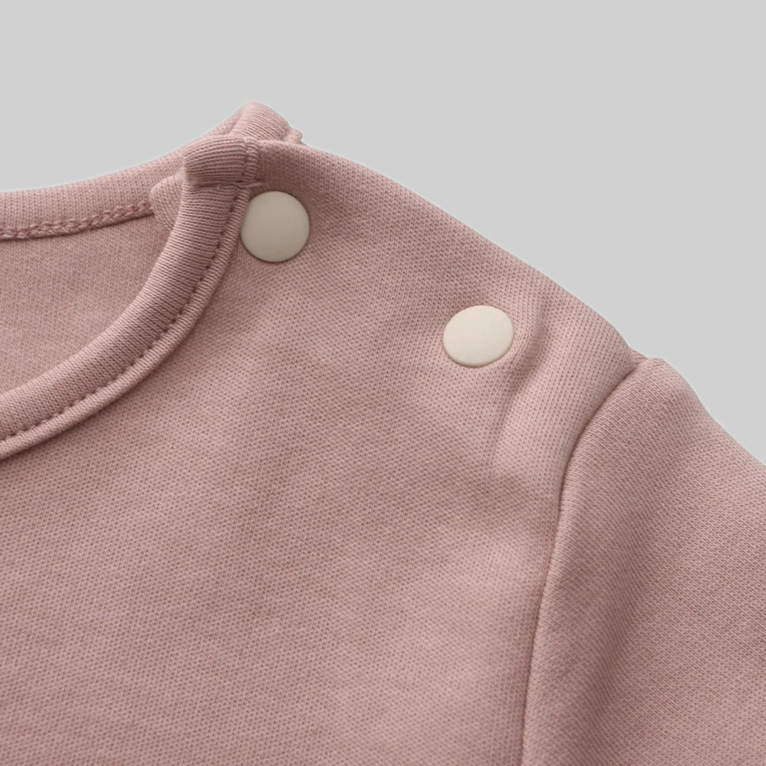 Close-up of a dusty pink baby romper showing the soft fabric, neckline stitching and two cream snap buttons at the shoulder.
