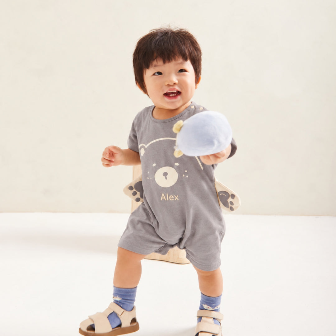 Smiling toddler wearing the personalised grey bear romper with the name “Alex,” holding a soft toy in a bright neutral studio setting.