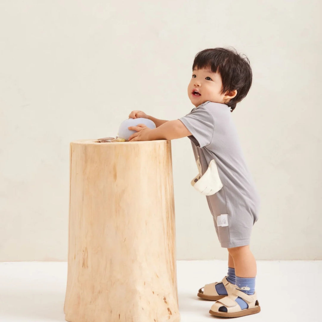 Toddler boy wearing the grey bear romper with 3D paw details, standing beside a wooden table in a neutral studio setting.