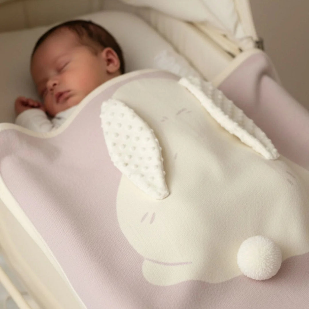 Sleeping baby tucked under a soft pink bunny blanket featuring a large white bunny applique with textured ears and pom-pom tail.
