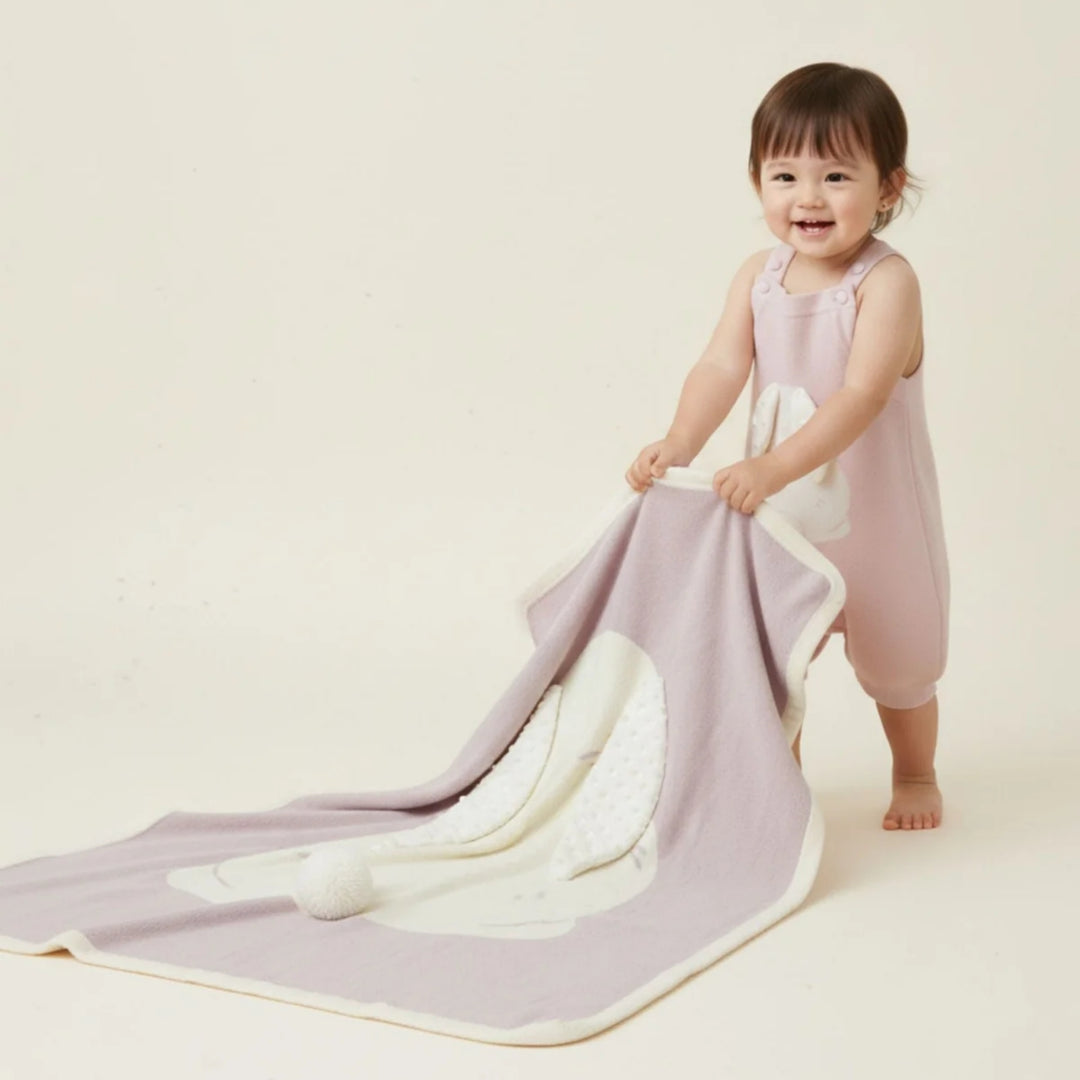 Smiling toddler in a pale pink outfit holding and pulling a soft pink bunny blanket with a large white bunny applique and textured ears.