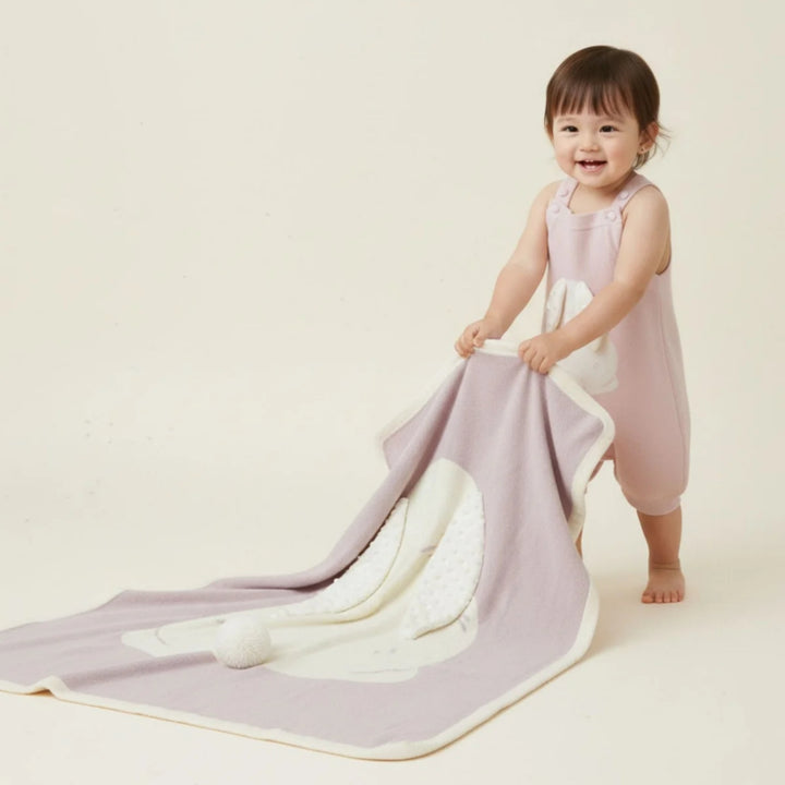 Smiling toddler in a pale pink outfit holding and pulling a soft pink bunny blanket with a large white bunny applique and textured ears.
