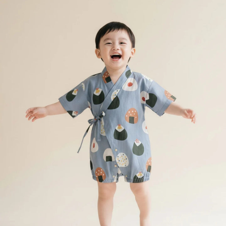 Young boy wearing a light blue onigiri-print kimono romper with wrap-front tie detail, standing and smiling against a neutral background.