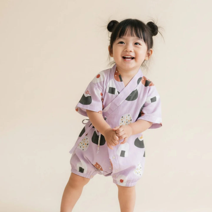 Toddler girl wearing a lilac onigiri-print kimono romper with wrap-front tie detail, standing and smiling against a soft neutral background.