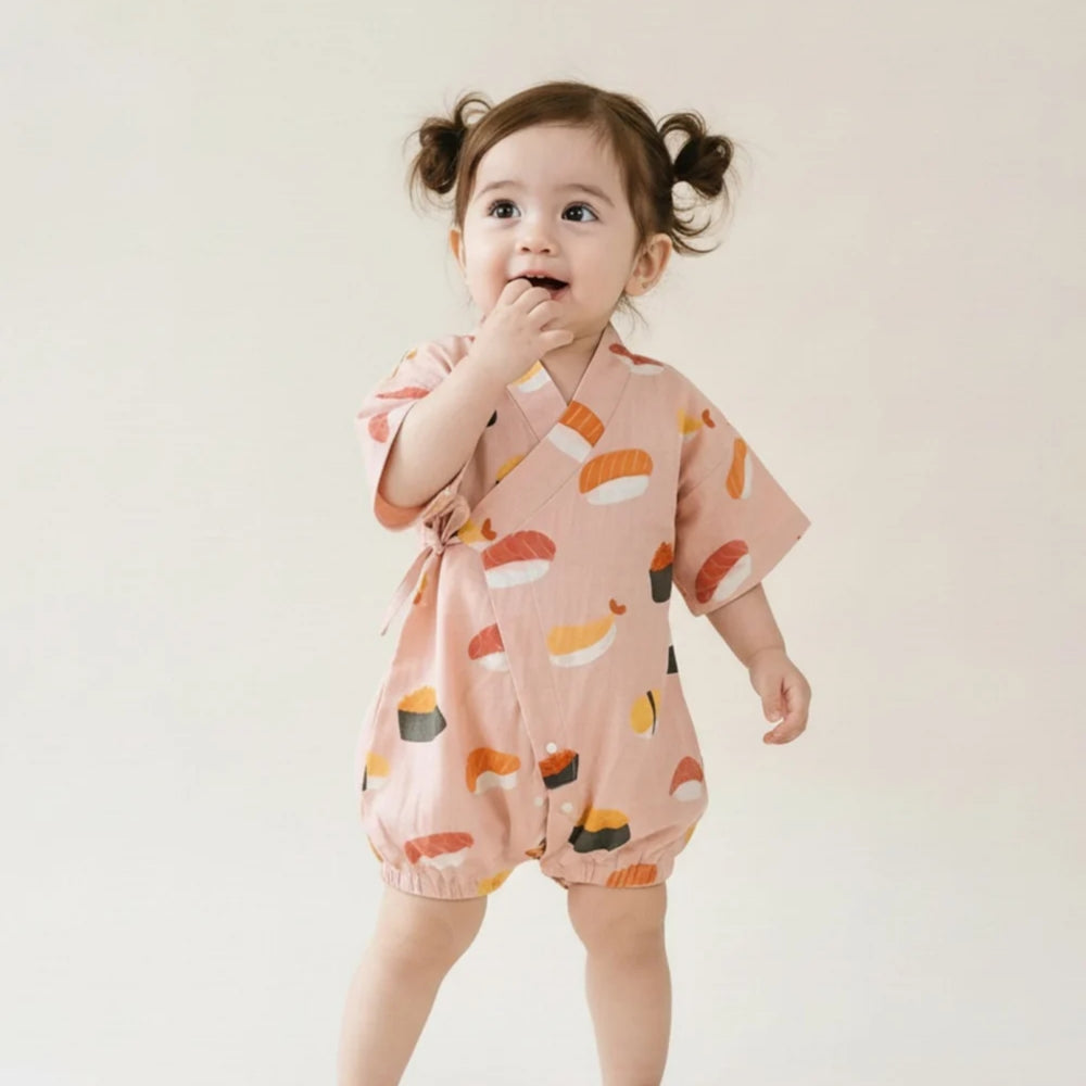 Toddler girl wearing a pink sushi-print kimono romper, standing against a soft neutral background.
