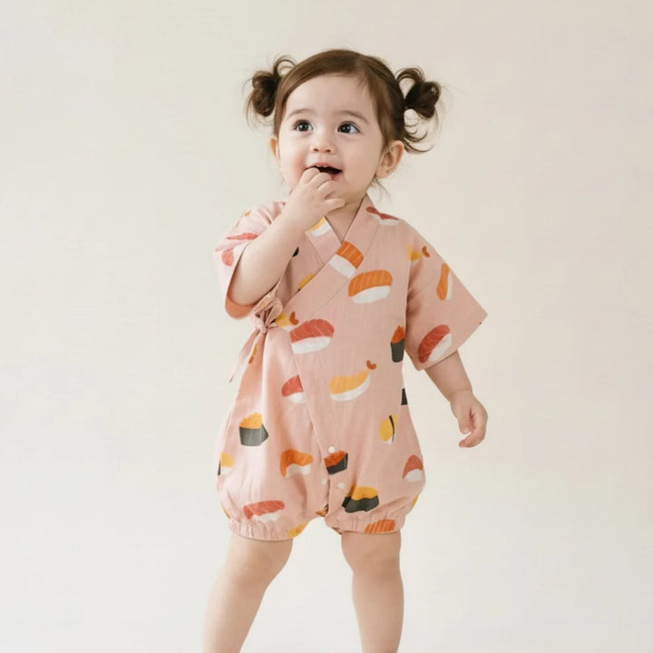 Toddler girl wearing a pink sushi-print kimono romper, standing against a soft neutral background.