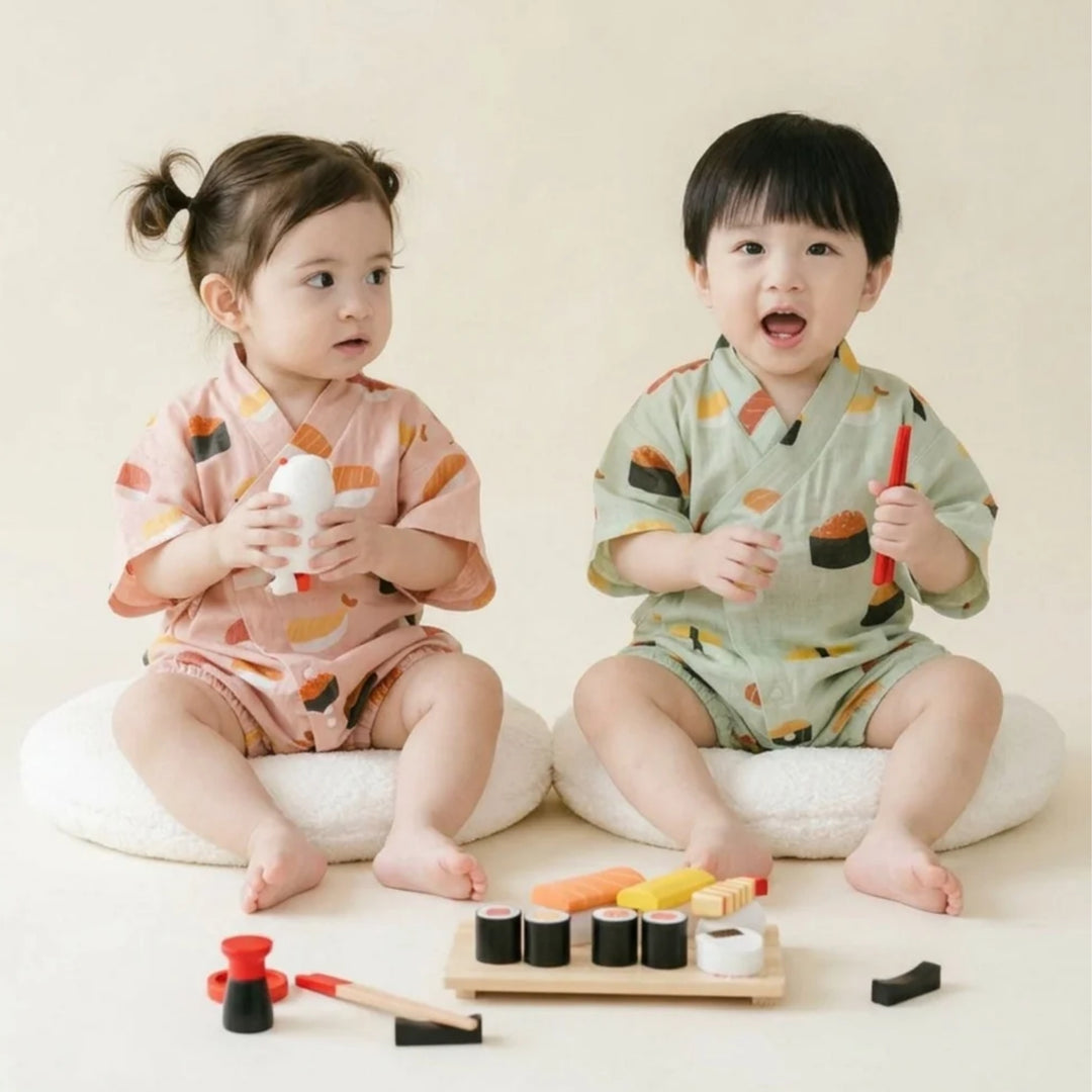 Two toddlers wearing matching sushi-print kimono rompers in pink and light green, sitting together with toy sushi pieces.