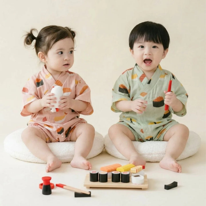 Two toddlers wearing matching sushi-print kimono rompers in pink and light green, sitting together with toy sushi pieces.