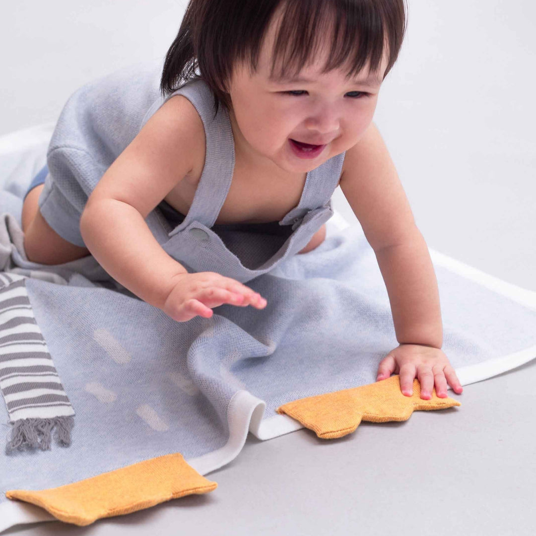 Smiling baby crawling on a soft light blue penguin blanket featuring an orange foot detail and grey-and-white striped scarf.