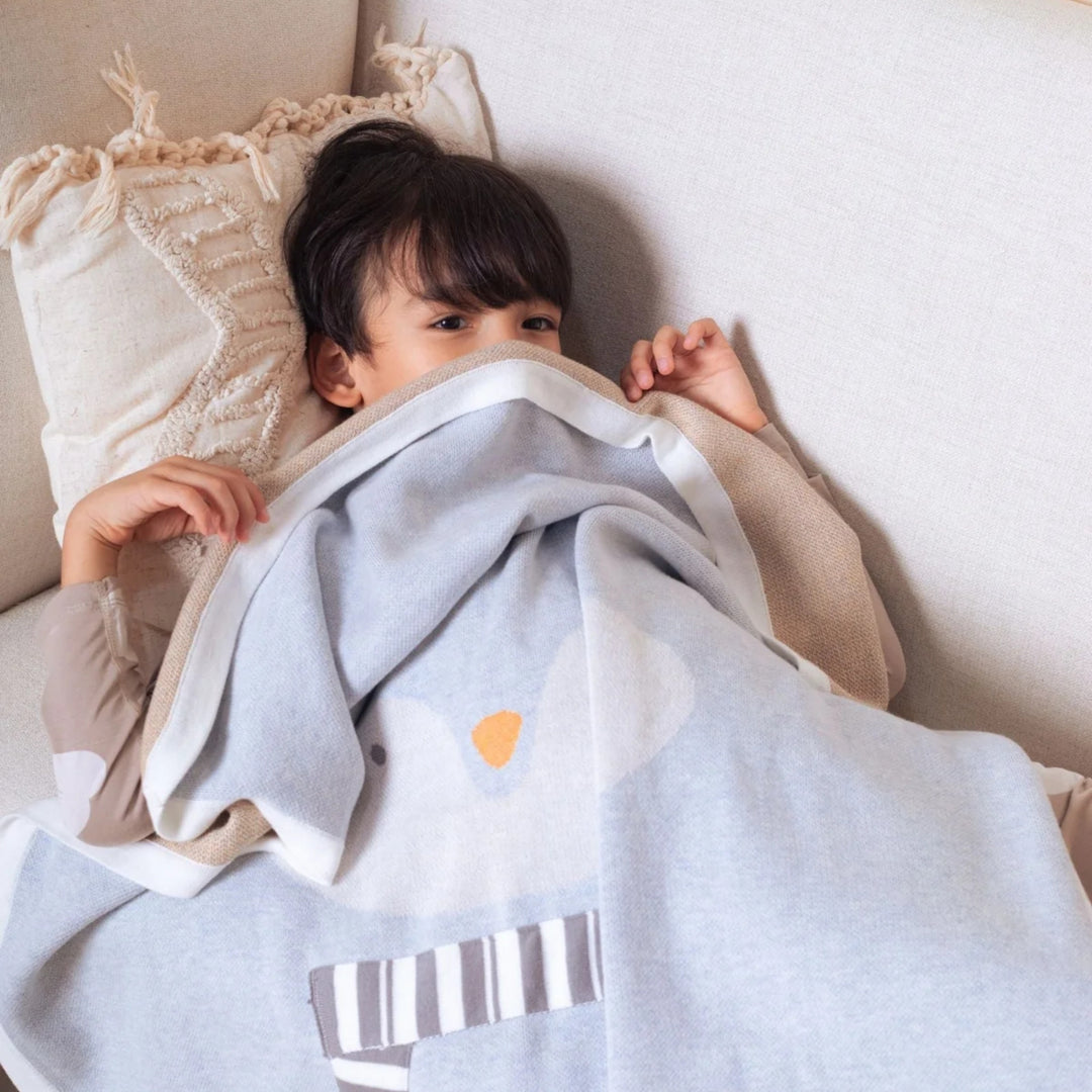 Young boy relaxing on a sofa under a light blue penguin blanket with white trim, orange beak detail, and grey-and-white striped scarf.