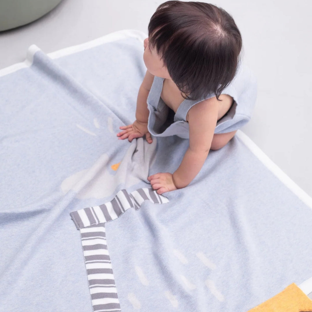 Baby sitting and playing on a light blue penguin blanket with white trim and grey-and-white striped scarf details.