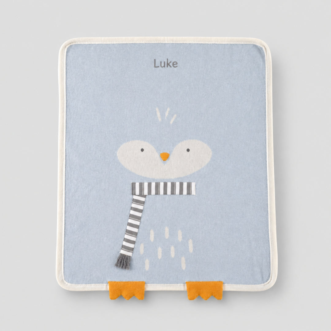 Light blue baby blanket with a white penguin face, orange beak and feet, grey-and-white striped scarf, and the name “Luke” embroidered at the top.