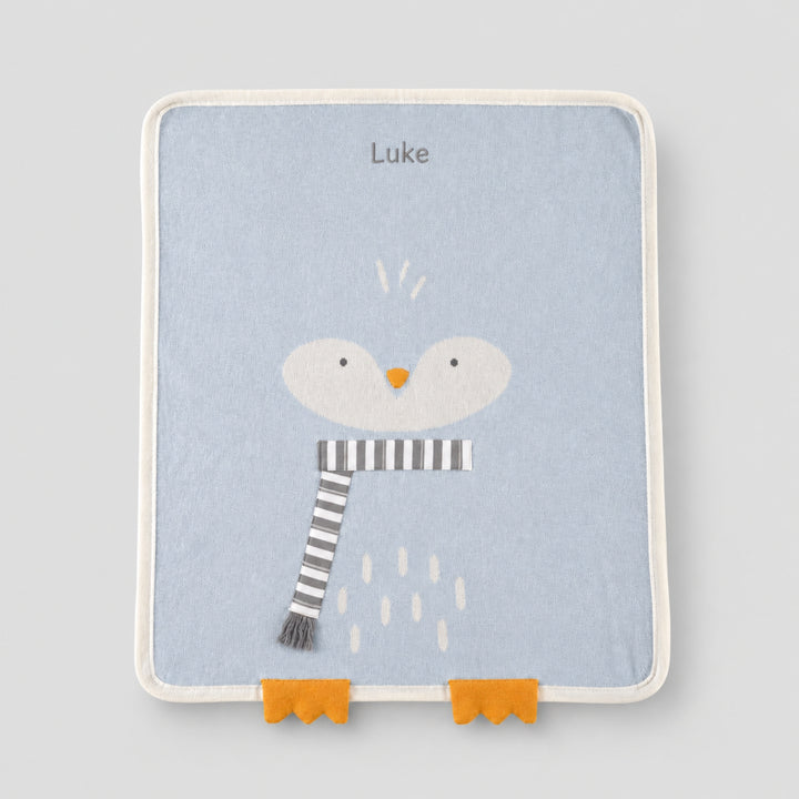 Light blue baby blanket with a white penguin face, orange beak and feet, grey-and-white striped scarf, and the name “Luke” embroidered at the top.