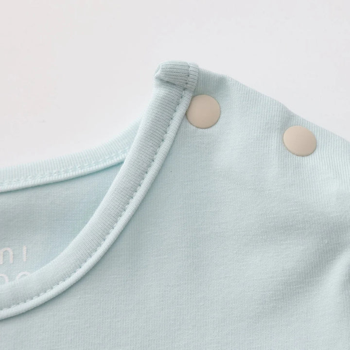 Close-up of the light blue romper showing the soft fabric, neckline stitching and two cream snap buttons at the shoulder.