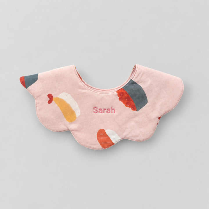 Pink scalloped baby bib with colourful sushi illustrations and the name “Sarah” embroidered at the centre.
