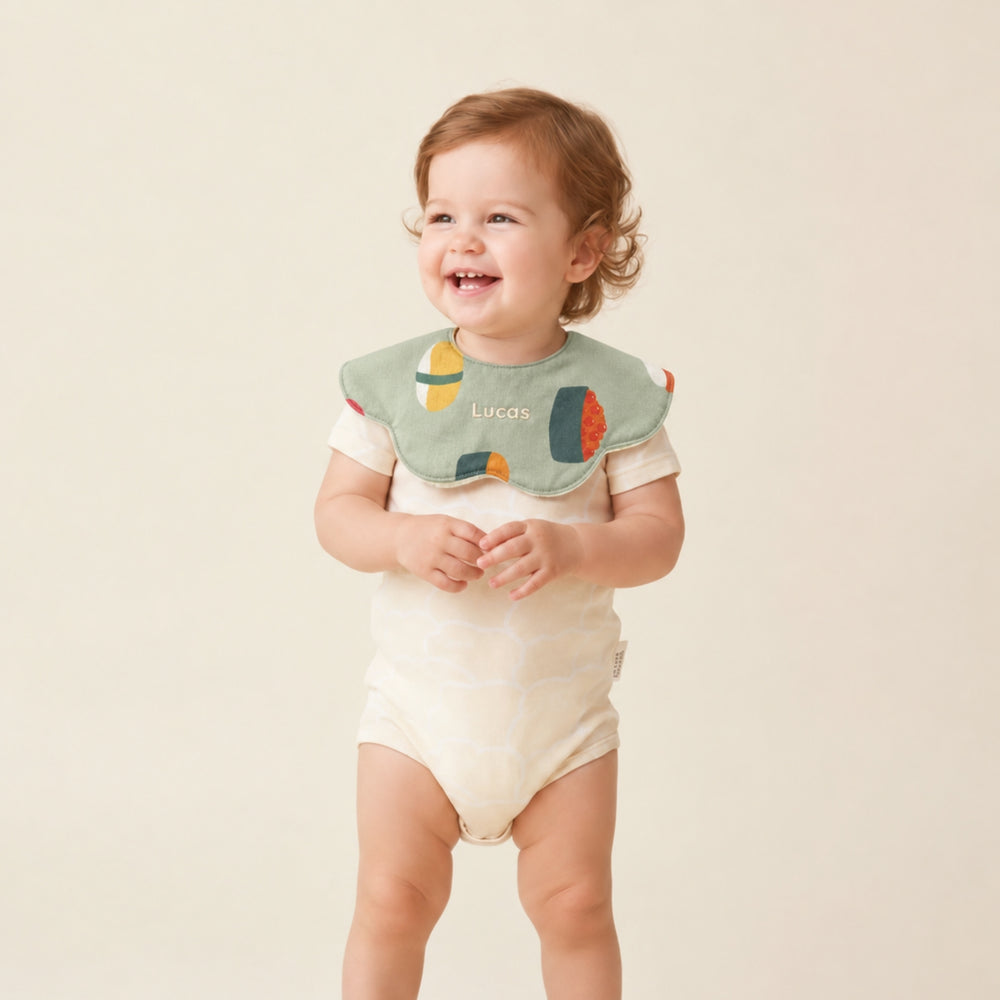 Smiling baby wearing a green sushi-print bib personalised with the name “Lucas,” paired with a cream bodysuit.