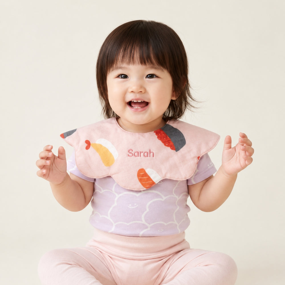 Smiling baby girl wearing a pink sushi-print bib personalised with the name “Sarah,” styled with a light purple top and pink bottoms.