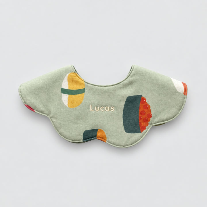 Green scalloped baby bib with colourful sushi illustrations and the name “Lucas” embroidered at the centre.