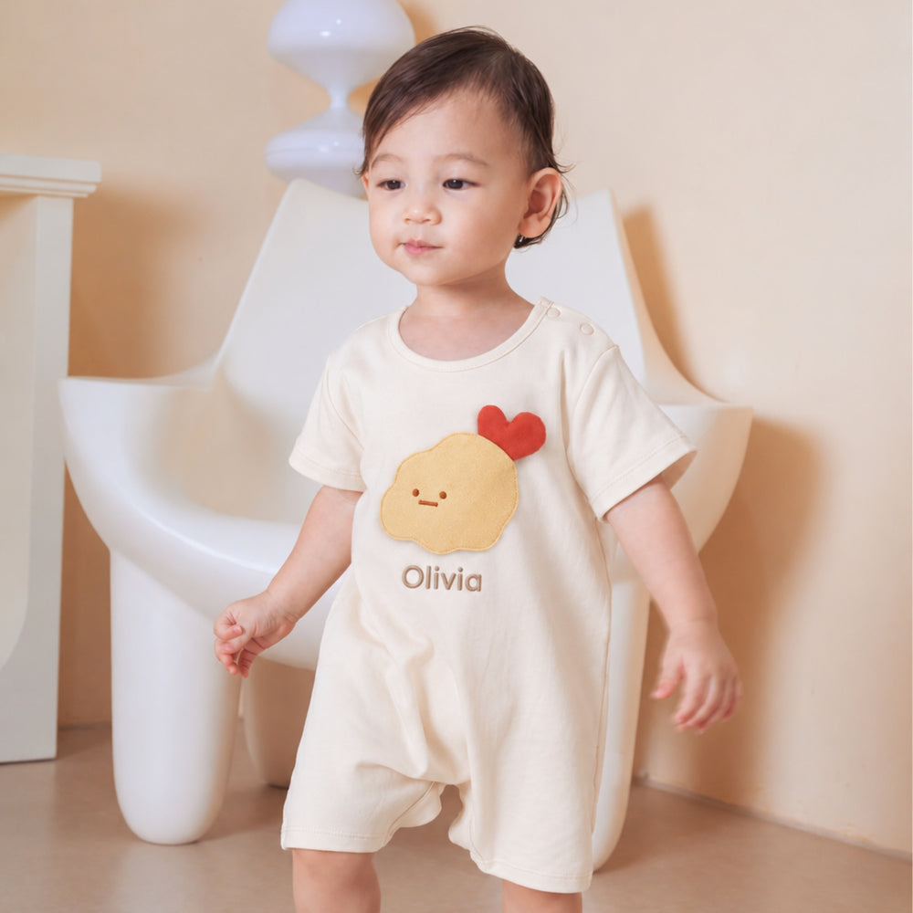 Toddler wearing a cream tempura romper personalised with the name “Olivia,” featuring a yellow tempura character and red heart detail.